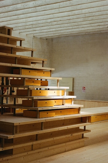 A geometric wooden structure composed of several stacked layers, with each layer featuring shelves or compartments. Some areas are illuminated by natural light, giving contrast to the warm tones of the wood. The setting is a modern, minimalist interior with a textured concrete ceiling.