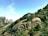 Rocky mountains covered with lush green trees and vegetation dominate the landscape. Peaks of the mountains rise dramatically in the background under a clear blue sky, with a few clouds floating gently above. A sense of tranquility and natural beauty is present.