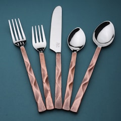 A set of cutlery consisting of two forks, a knife, and two spoons arranged vertically on a teal background. The handles have a copper tone with a textured, faceted design, while the fork and spoon heads, including the knife blade, are polished, reflecting light.
