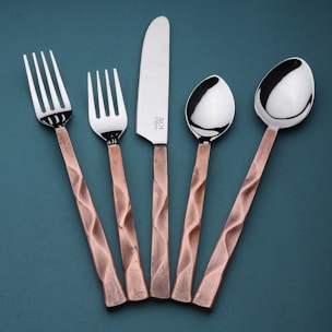 A set of cutlery consisting of two forks, a knife, and two spoons arranged vertically on a teal background. The handles have a copper tone with a textured, faceted design, while the fork and spoon heads, including the knife blade, are polished, reflecting light.