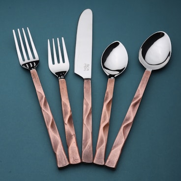 A set of cutlery consisting of two forks, a knife, and two spoons arranged vertically on a teal background. The handles have a copper tone with a textured, faceted design, while the fork and spoon heads, including the knife blade, are polished, reflecting light.