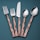 A set of cutlery consisting of two forks, a knife, and two spoons arranged vertically on a teal background. The handles have a copper tone with a textured, faceted design, while the fork and spoon heads, including the knife blade, are polished, reflecting light.