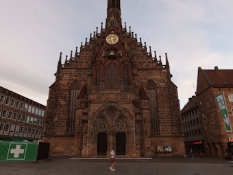 A large, ornate Gothic-style church with a prominent clock tower, intricate stone carvings, and arched windows. A person can be seen walking in front of the building. Surrounding the church are modern buildings, including a green tent with a medical cross symbol.
