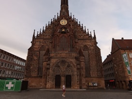 A large, ornate Gothic-style church with a prominent clock tower, intricate stone carvings, and arched windows. A person can be seen walking in front of the building. Surrounding the church are modern buildings, including a green tent with a medical cross symbol.