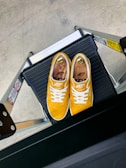 A pair of vibrant yellow sneakers with white laces positioned neatly on a black step ladder. The sneakers have wooden shoe trees inside and a logo that reads 'SNEEP CREW' is visible. The step ladder is metallic with black and silver components, placed on a light gray concrete floor.