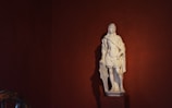 A classical white marble statue depicting a male figure draped in traditional Roman attire stands against a dark red background. The figure holds a shield by his side, exuding a strong and heroic presence.