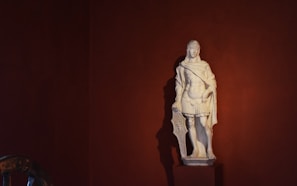 A classical white marble statue depicting a male figure draped in traditional Roman attire stands against a dark red background. The figure holds a shield by his side, exuding a strong and heroic presence.