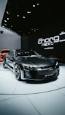 A sleek, modern electric car with a dark finish is displayed on a white platform in an indoor showroom. The setting features upscale lighting that highlights the car's design. In the background, branding elements and text like 'the charge next' are visible, along with another vehicle partially visible.