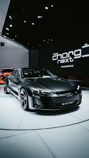 A sleek, modern electric car with a dark finish is displayed on a white platform in an indoor showroom. The setting features upscale lighting that highlights the car's design. In the background, branding elements and text like 'the charge next' are visible, along with another vehicle partially visible.