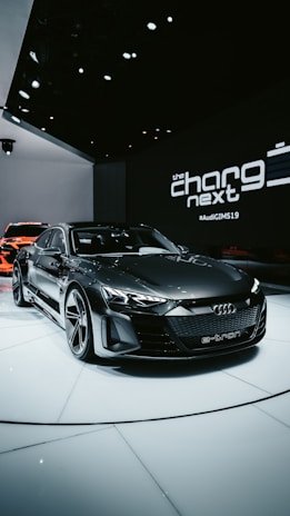 A sleek, modern electric car with a dark finish is displayed on a white platform in an indoor showroom. The setting features upscale lighting that highlights the car's design. In the background, branding elements and text like 'the charge next' are visible, along with another vehicle partially visible.