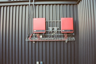 Two red electrical devices are mounted on a black corrugated metal wall, connected by a series of cables. The setup includes several black wires attached to the devices and a metal rack supporting the components.