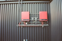 Two red electrical devices are mounted on a black corrugated metal wall, connected by a series of cables. The setup includes several black wires attached to the devices and a metal rack supporting the components.