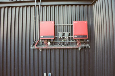 Two red electrical devices are mounted on a black corrugated metal wall, connected by a series of cables. The setup includes several black wires attached to the devices and a metal rack supporting the components.