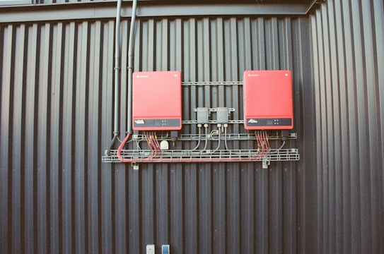 Two red electrical devices are mounted on a black corrugated metal wall, connected by a series of cables. The setup includes several black wires attached to the devices and a metal rack supporting the components.