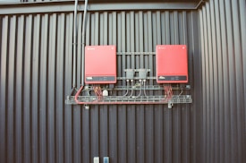 Two red electrical devices are mounted on a black corrugated metal wall, connected by a series of cables. The setup includes several black wires attached to the devices and a metal rack supporting the components.