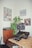 A room corner features a wooden cabinet topped with a turntable and a speaker. A large speaker with two smaller electronic devices and a yellow cable lies on a wooden desk. A leafy plant and wall art including posters and an album cover add character to the space.