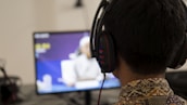 A person wearing headphones is seated in front of a computer monitor. The screen displays blurred content, possibly a video or a game. The person has short hair and is wearing a patterned shirt.