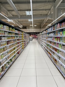 A supermarket aisle lined with neatly organized shelves filled with various packaged goods. The shelves display an assortment of products on both sides, extending into the background. Bright lighting from the ceiling illuminates the space, and a tiled floor is visible. Signage in the distance indicates different sections of the store.