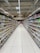 A supermarket aisle lined with neatly organized shelves filled with various packaged goods. The shelves display an assortment of products on both sides, extending into the background. Bright lighting from the ceiling illuminates the space, and a tiled floor is visible. Signage in the distance indicates different sections of the store.