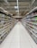 A supermarket aisle lined with neatly organized shelves filled with various packaged goods. The shelves display an assortment of products on both sides, extending into the background. Bright lighting from the ceiling illuminates the space, and a tiled floor is visible. Signage in the distance indicates different sections of the store.