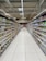 A supermarket aisle lined with neatly organized shelves filled with various packaged goods. The shelves display an assortment of products on both sides, extending into the background. Bright lighting from the ceiling illuminates the space, and a tiled floor is visible. Signage in the distance indicates different sections of the store.