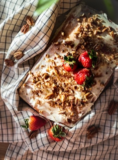 A homemade cake topped with cream frosting, pecans, and fresh strawberries, placed on a checkered cloth with additional strawberries and pecans scattered around.