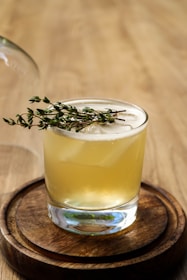 A glass of pale yellow beverage is set on a wooden coaster. The drink is garnished with a sprig of fresh herbs, possibly thyme. The background is a smooth, light wooden surface, creating a warm and inviting atmosphere.