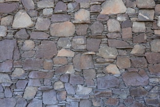 A wall constructed of irregularly shaped, stacked stone slabs with a mix of earthy tones and textures. The stones vary in size, from large flat pieces to smaller ones, and are meticulously arranged to fit together tightly.