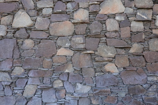 A wall constructed of irregularly shaped, stacked stone slabs with a mix of earthy tones and textures. The stones vary in size, from large flat pieces to smaller ones, and are meticulously arranged to fit together tightly.