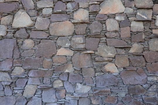 A wall constructed of irregularly shaped, stacked stone slabs with a mix of earthy tones and textures. The stones vary in size, from large flat pieces to smaller ones, and are meticulously arranged to fit together tightly.