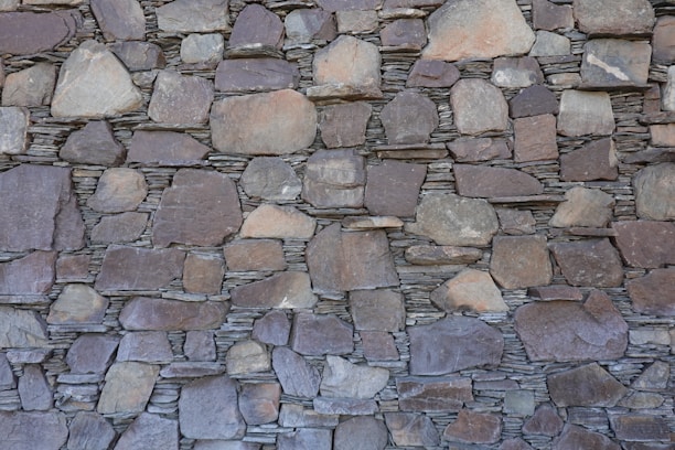 A wall constructed of irregularly shaped, stacked stone slabs with a mix of earthy tones and textures. The stones vary in size, from large flat pieces to smaller ones, and are meticulously arranged to fit together tightly.