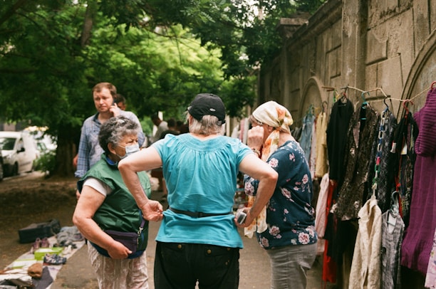 A small group of people is gathered at an outdoor market or street vendor, with a focus on a woman in a blue shirt and black cap who is interacting with another woman in a floral shirt with a headscarf. There is a clothing rack to the right with various garments hanging. Trees and a stone wall with arches provide the backdrop, while another man walks by in the background, talking on a phone.