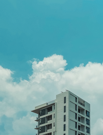 A tall, modern apartment building with multiple balconies is set against a bright blue sky filled with fluffy white clouds.