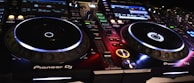 A DJ controller setup with illuminated buttons and displays, showcasing various controls and interfaces. The equipment is branded with Pioneer DJ, featuring two turntables and multiple digital screens displaying track information.