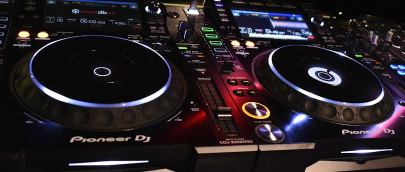A DJ controller setup with illuminated buttons and displays, showcasing various controls and interfaces. The equipment is branded with Pioneer DJ, featuring two turntables and multiple digital screens displaying track information.