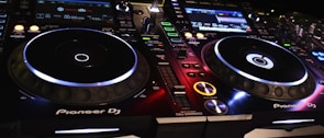 A DJ controller setup with illuminated buttons and displays, showcasing various controls and interfaces. The equipment is branded with Pioneer DJ, featuring two turntables and multiple digital screens displaying track information.