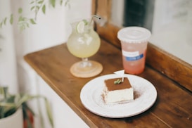 A wooden table holds a plate with a piece of tiramisu garnished with a small flag and a sprig of mint. Next to it, there is a glass of lime beverage with a straw and a slice of lime on the rim, placed on a coaster. A disposable cup with a lid and drink beside the glass adds to the scene. The setup is near a window, allowing natural light to softly illuminate the objects, and some leaves are visible in the corner, suggesting the presence of plants.