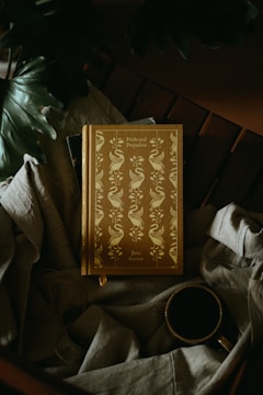 A hardcover book with an ornate cover featuring a swan and floral pattern design rests on a fabric surface next to a cup of coffee. The book's title, Pride and Prejudice by Jane Austen, is prominently visible. The setting includes large green leaves partially visible in the upper left corner, adding a natural touch.