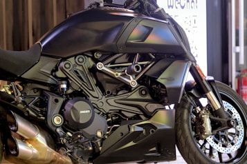 A sleek, modern motorcycle with a matte black finish is prominently displayed. The close-up view highlights the intricate details of its engine and bodywork, showcasing the brand logo on the engine cover. The vehicle's design features sharp lines and polished metallic elements, with dual exhaust pipes visible at the bottom.