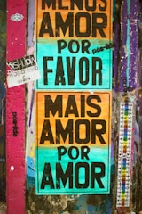 A wall covered with colorful graffiti and posters. Two prominent posters with large black text on multicolored backgrounds. One reads 'Menos Amor Por Favor' and the other 'Mais Amor Por Amor'. The wall also has various other small stickers and graffiti in different colors, mainly purple and teal.