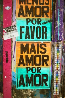 A wall covered with colorful graffiti and posters. Two prominent posters with large black text on multicolored backgrounds. One reads 'Menos Amor Por Favor' and the other 'Mais Amor Por Amor'. The wall also has various other small stickers and graffiti in different colors, mainly purple and teal.
