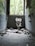 A dilapidated bathroom with a broken toilet and scattered debris on the floor. The walls are tiled in blue and covered with graffiti. A large window at the far end allows natural light to illuminate the room, revealing overgrown vegetation outside.