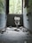A dilapidated bathroom with a broken toilet and scattered debris on the floor. The walls are tiled in blue and covered with graffiti. A large window at the far end allows natural light to illuminate the room, revealing overgrown vegetation outside.