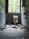 A dilapidated bathroom with a broken toilet and scattered debris on the floor. The walls are tiled in blue and covered with graffiti. A large window at the far end allows natural light to illuminate the room, revealing overgrown vegetation outside.