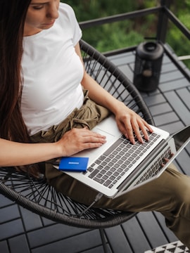 A person seated on a balcony chair, typing on a laptop with a Samsung external hard drive connected. The setting appears relaxed, with a black lantern placed on the table beside. The person is wearing a white t-shirt and khaki pants.