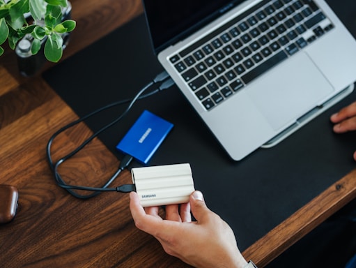 A person holds a white Samsung portable SSD connected to a laptop on a wooden desk. A blue Samsung external hard drive rests nearby, and a potted plant is visible on the desk.