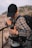 A person wearing a black cap and a plaid shirt is standing outdoors, adjusting or examining an item from their belt or backpack. The person is carrying a large backpack, and there is a carabiner attached to their outfit. The background features a blurry landscape of rocky hills or mountains.