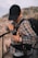 A person wearing a black cap and a plaid shirt is standing outdoors, adjusting or examining an item from their belt or backpack. The person is carrying a large backpack, and there is a carabiner attached to their outfit. The background features a blurry landscape of rocky hills or mountains.