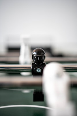 A close-up view of a foosball table, focusing on a black foosball player figure mounted on a metal rod. The background is out of focus, making the figure the central element in the image.