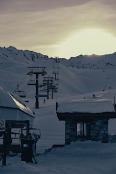 A ski resort scene at dusk with ski lifts ascending a snow-covered mountain slope. The sun is setting behind the mountains, casting a dim light over the landscape. There are a few people skiing down the slopes, and a small building with a snow-covered roof is visible in the foreground.